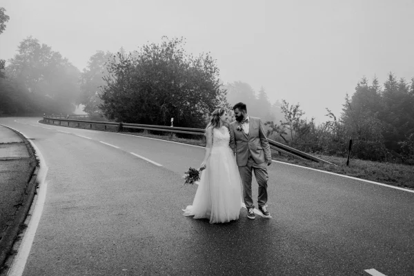 Brautpaar spaziert Hand in Hand auf einer nebligen Landstraße, eingefangen in einer stimmungsvollen Schwarz-Weiß-Aufnahme