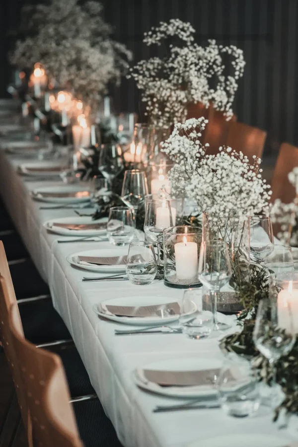 Festlich gedeckte Hochzeitstafel mit weißen Tellern, Weingläsern und Kerzen. Die Dekoration besteht aus Schleierkraut und grünen Zweigen, die das warme Kerzenlicht reflektieren.