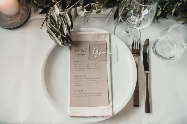 Detailaufnahme eines Hochzeitstisches mit weißem Teller, Besteck und einem eleganten Menükärtchen mit der Aufschrift „Groom“. Dekoriert mit Olivenzweigen und einer Kerze im Glas.