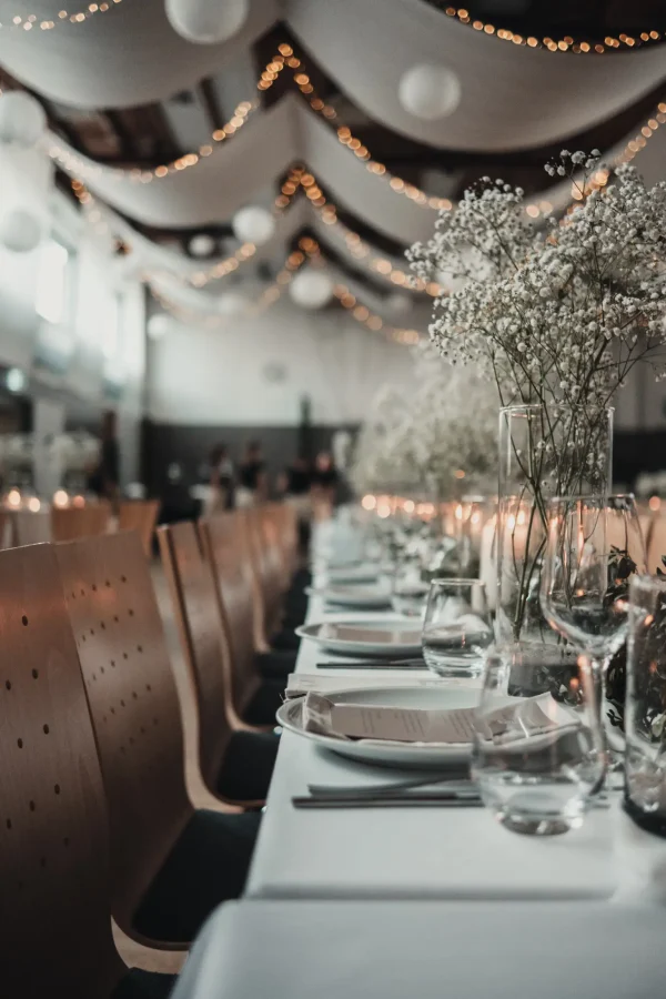 Lange, festlich gedeckte Hochzeitstafel in einer dekorierten Halle mit hängenden Stoffbahnen und Lichterketten. Die Tischdekoration besteht aus Schleierkraut, Kerzen und elegantem Geschirr.