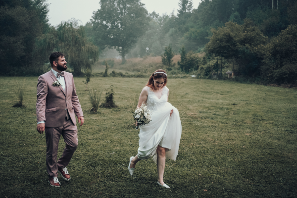 Braut hebt ihr Kleid an und läuft lächelnd über eine feuchte Wiese, Bräutigam folgt