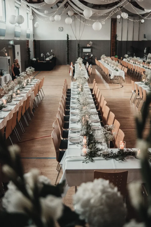 festlich gedeckte Hochzeitstafel in einer dekorierten Halle. Die Tische sind mit Schleierkraut, Olivenzweigen und Kerzen geschmückt, während Lichterketten und Stoffbahnen von der Decke hängen.