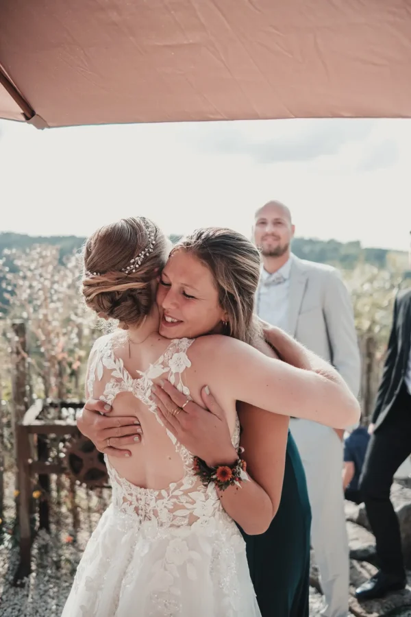 Braut in einem detailliert bestickten Kleid umarmt herzlich eine Freundin unter einem Sonnenschirm, während im Hintergrund Gäste lächelnd zuschauen. Die Szene spielt sich auf einer sonnendurchfluteten Terrasse mit malerischer Aussicht ab.