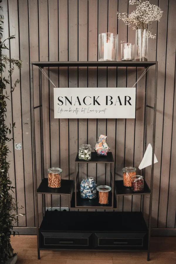 Hochzeits-Snackbar mit verschiedenen Snacks in Gläsern, dekoriert mit einem Schild „Snack Bar – Life can be salty, but love is sweet“, Kerzen und Schleierkraut auf einem Metallregal.