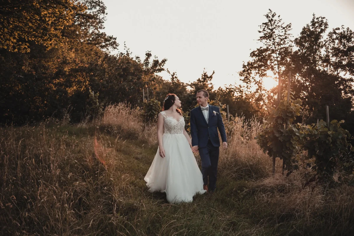 Das Brautpaar spaziert Hand in Hand durch die goldenen Weinberge der Heuchelberger Warte. Die warme Abendsonne taucht die Szene in ein traumhaftes Licht, während sie sich verliebt anblicken.