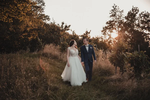 Das Brautpaar spaziert Hand in Hand durch die goldenen Weinberge der Heuchelberger Warte. Die warme Abendsonne taucht die Szene in ein traumhaftes Licht, während sie sich verliebt anblicken.