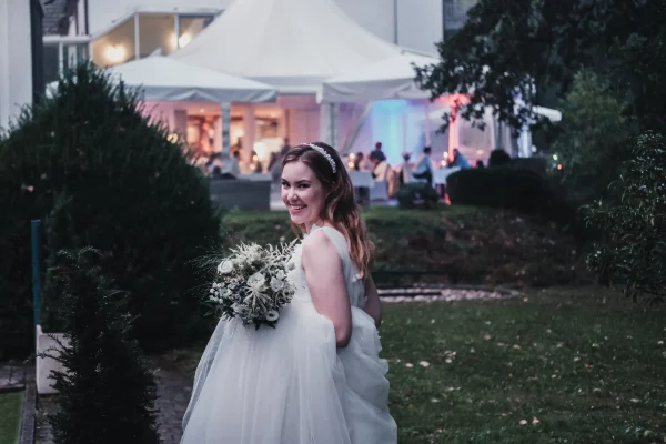 Braut mit Blumenstrauß blickt lächelnd über die Schulter in Richtung Festzelt