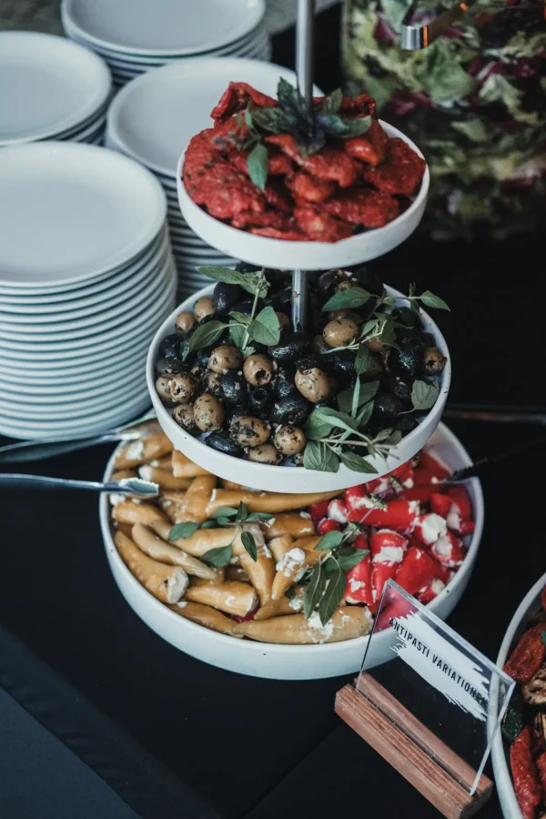 Dreistöckige Antipasti-Etagere mit gefüllten Peperoni, Oliven und getrockneten Tomaten, dekoriert mit frischem Basilikum. Daneben gestapelte Teller für die Gäste.
