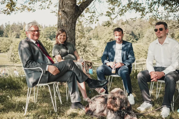 Hochzeitsgäste auf gemütlichen Stühlen unter einer schattigen Linde am Nachmittag. Hund auf der Wiese in der Mitte der Gruppe.