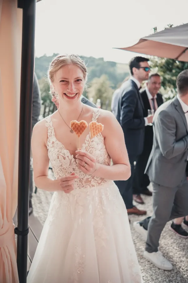 Die Braut lächelnd beim Sektempfang mit zwei Herzwaffeln am Holzstiel