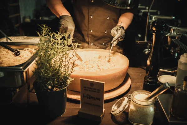Live-Cooking-Station bei der Hochzeit: Ein Koch bereitet frische Pasta in einem großen Parmesanlaib zu. Kräuter, Parmesan und Pfeffermühle als Dekoration auf dem Tisch.