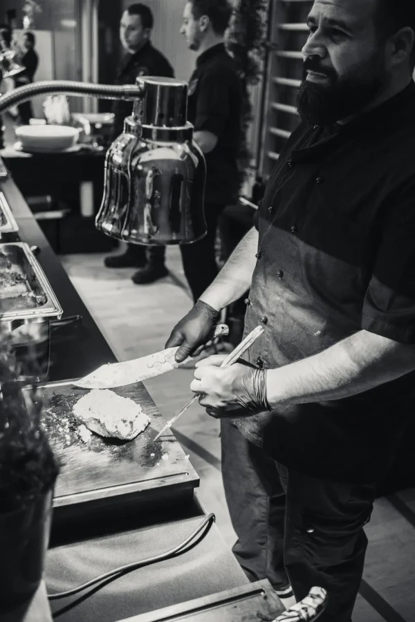 Schwarz-Weiß-Fotografie eines Kochs an der Live-Cooking-Station, der ein Stück Fleisch mit einem Messer und einer Zange zubereitet. Im Hintergrund stehen weitere Köche bereit.