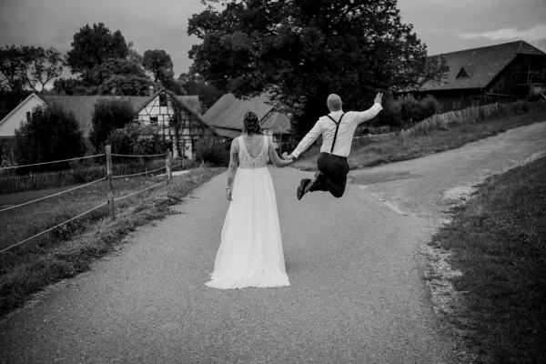 Ansicht des Brautpaars von hinten. Location im Hintergrund. Der Bräutigam hält seine Frau an der Hand und vollführt einen ausgelassen Luftsprung vor Freude.