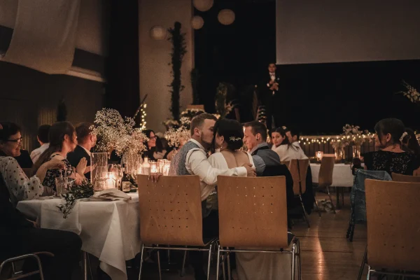 Romantischer Moment während der Hochzeitsfeier: Der Bräutigam gibt der Braut einen Kuss, während sie gemeinsam an einem festlich dekorierten Tisch mit Kerzen und Schleierkraut sitzen. Im Hintergrund hält ein Gast eine Rede auf der Bühne.