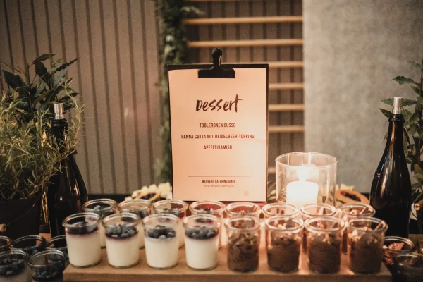 Dessertbuffet mit Gläsern gefüllt mit Tobleronemousse, Panna Cotta mit Heidelbeer-Topping und Apfeltiramisu. Im Hintergrund ein beleuchtetes Schild mit der Dessertauswahl, umgeben von Kräutern und Kerzen.