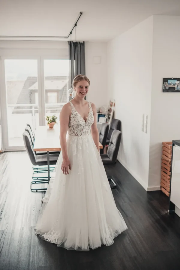 Die Braut in ihrem wunderschönen, weißen Hochzeitskleid mit floralen Spitzenapplikationen, strahlend im modernen Ambiente ihres Zuhauses.