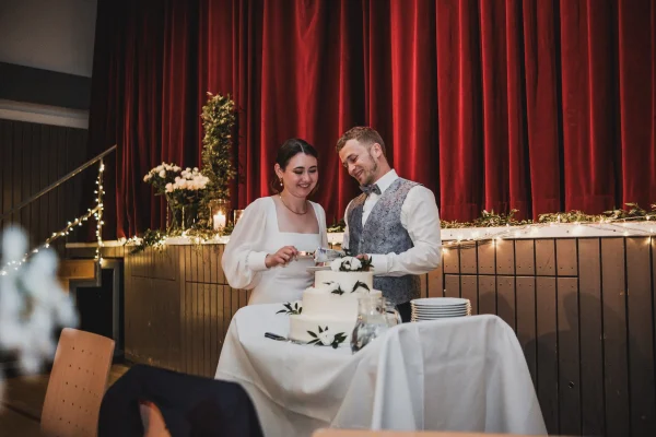 Das Brautpaar schneidet gemeinsam die Hochzeitstorte an, eine dreistöckige, weiße Torte mit Blumenverzierung. Im Hintergrund eine festlich geschmückte Bühne mit rotem Vorhang und Lichterketten.
