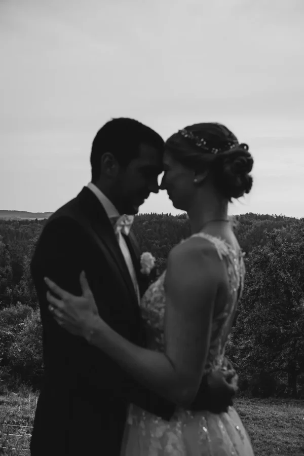 Schwarz-Weiß Aufnahme des Brautpaars, das in einer Umarmung Stirn an Stirn steht. Fokus auf den Wald hinter dem Paar.