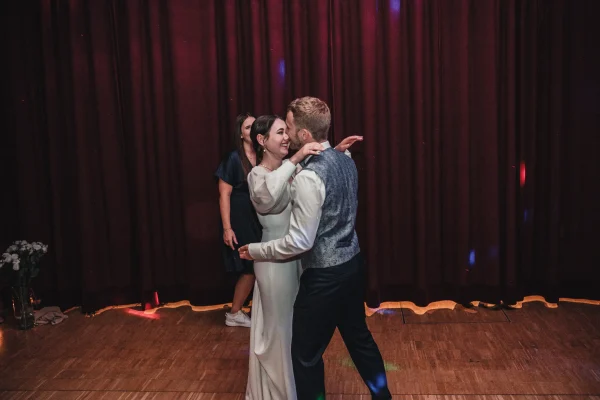 Romantischer Tanz des Brautpaares vor einem roten Vorhang. Die Braut lächelt den Bräutigam verliebt an, während sie sich in den Armen halten.