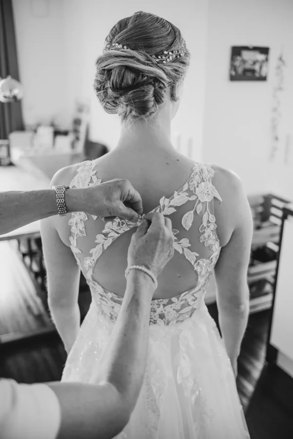 Detailaufnahme der Braut, wie die Knöpfe ihres Brautkleides von helfenden Händen verschlossen werden – ein Moment der Intimität und Liebe.