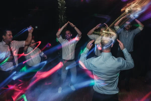 Hochzeitsgäste tanzen ausgelassen auf der Tanzfläche unter bunten Lichtern. Langzeitbelichtung fängt die Bewegung der Lichter ein und erzeugt eine dynamische Atmosphäre.
